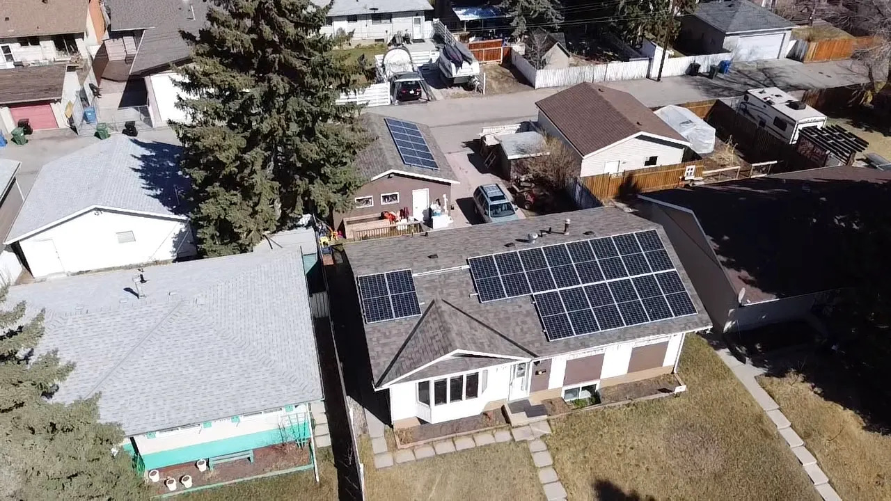 An aerial view of a residential neighborhood showing houses with solar panels on their roofs, driveways with vehicles, and a tree casting a shadow.