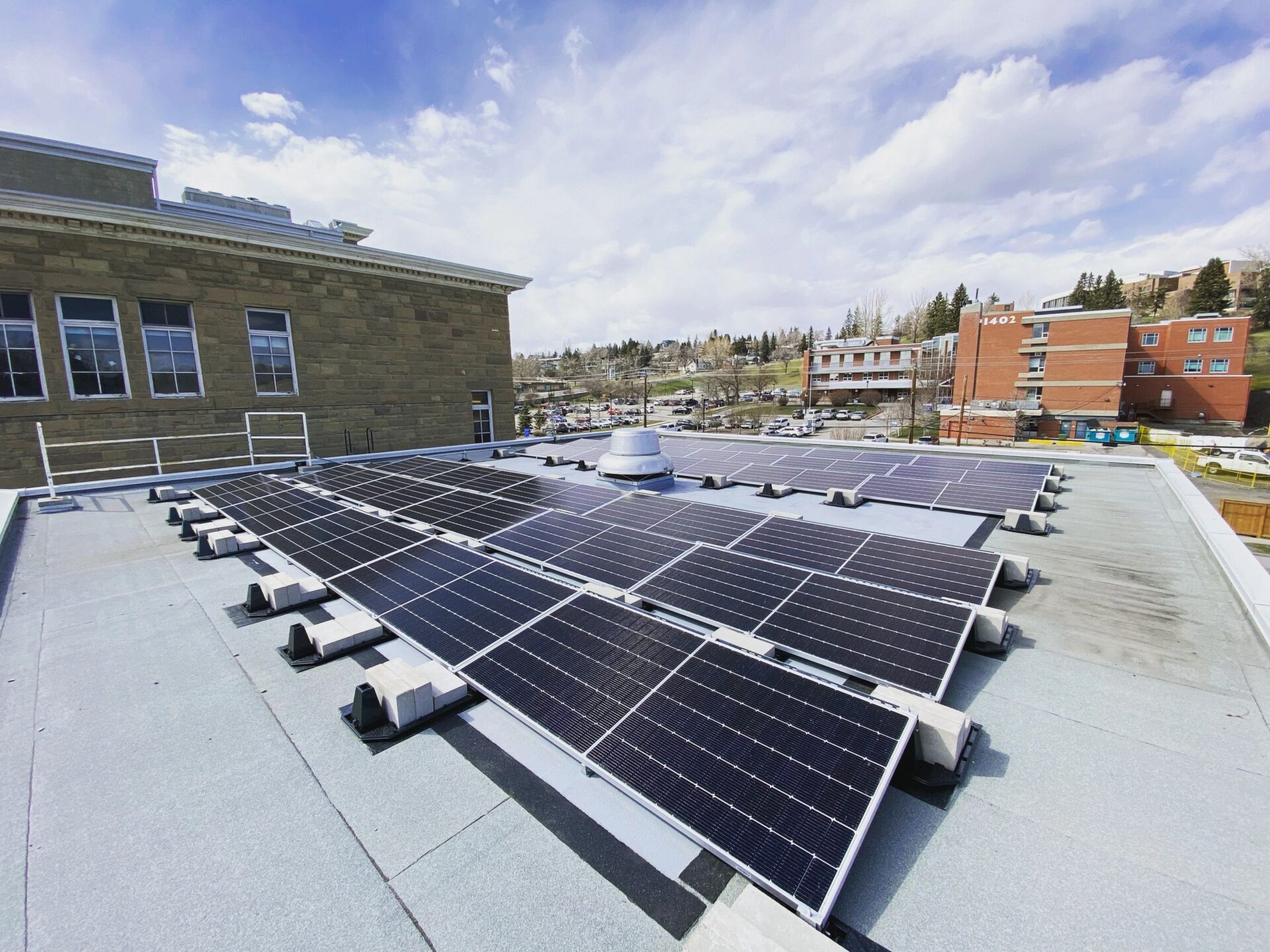 Aerial view of a commercial flat-roof solar panel installation with the Calgary city skyline and urban buildings in the background.