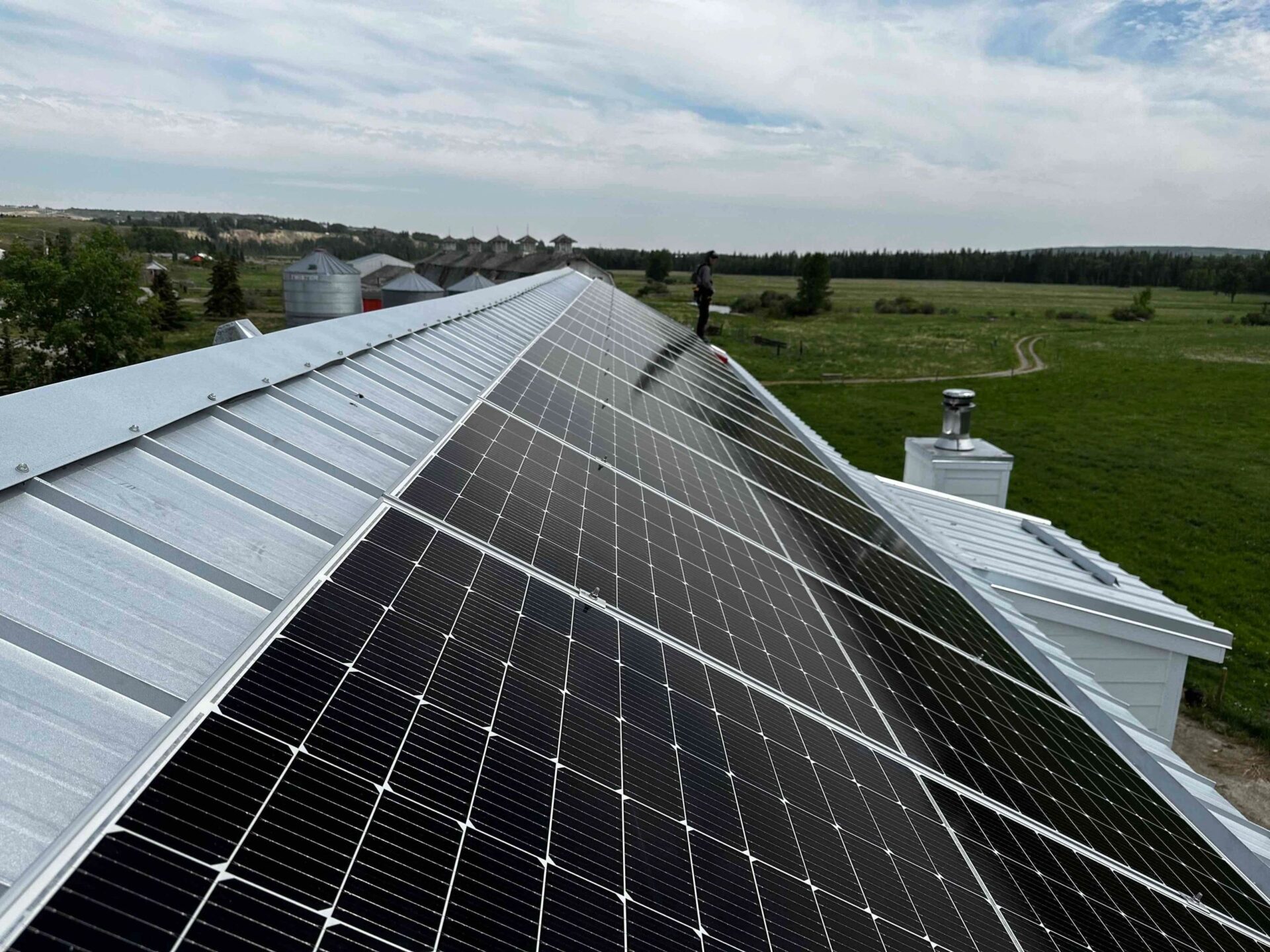 The image shows an array of solar panels installed on the sloping roof of a building, with rural landscape and silos in the background.