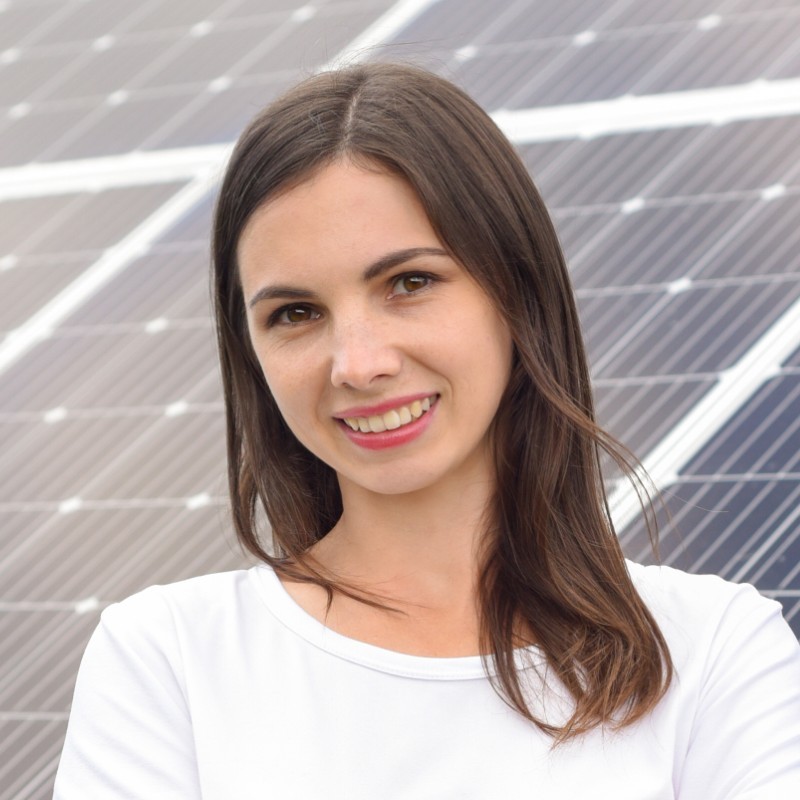 This is a portrait of a smiling person with shoulder-length brown hair, wearing a black top with a crisscross design, set against a gray backdrop.