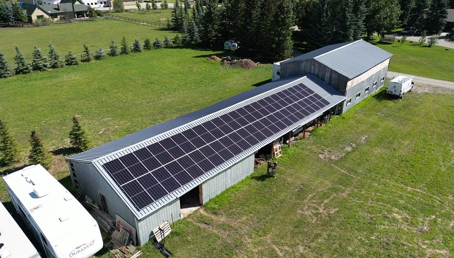 Aerial view of a rural property showing a large barn with a solar panel-covered roof, an RV, green grass, and a row of young trees.