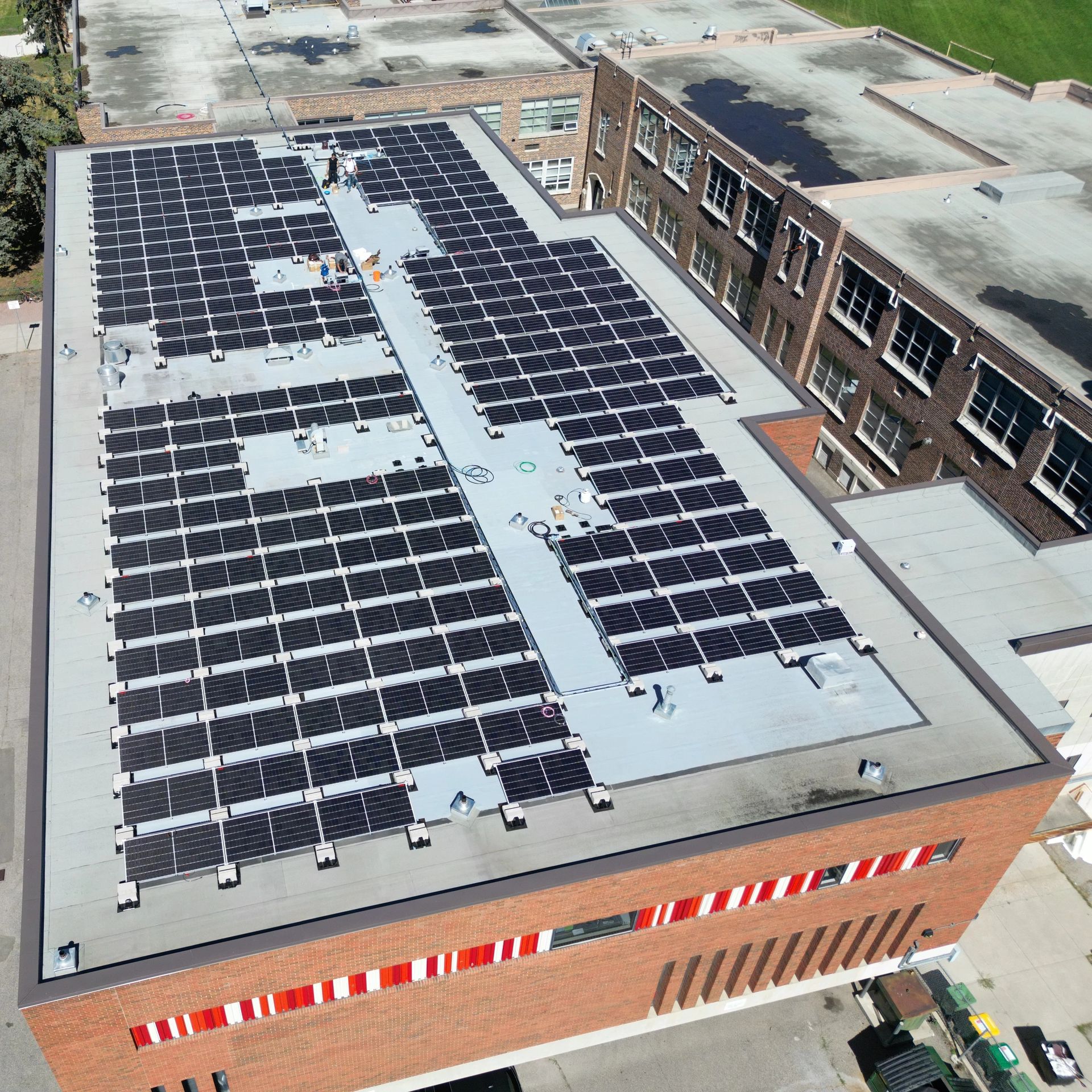 An aerial view of a large building with a flat roof densely covered in solar panels, indicating a focus on renewable energy. The surrounding area includes parked cars and greenery.
