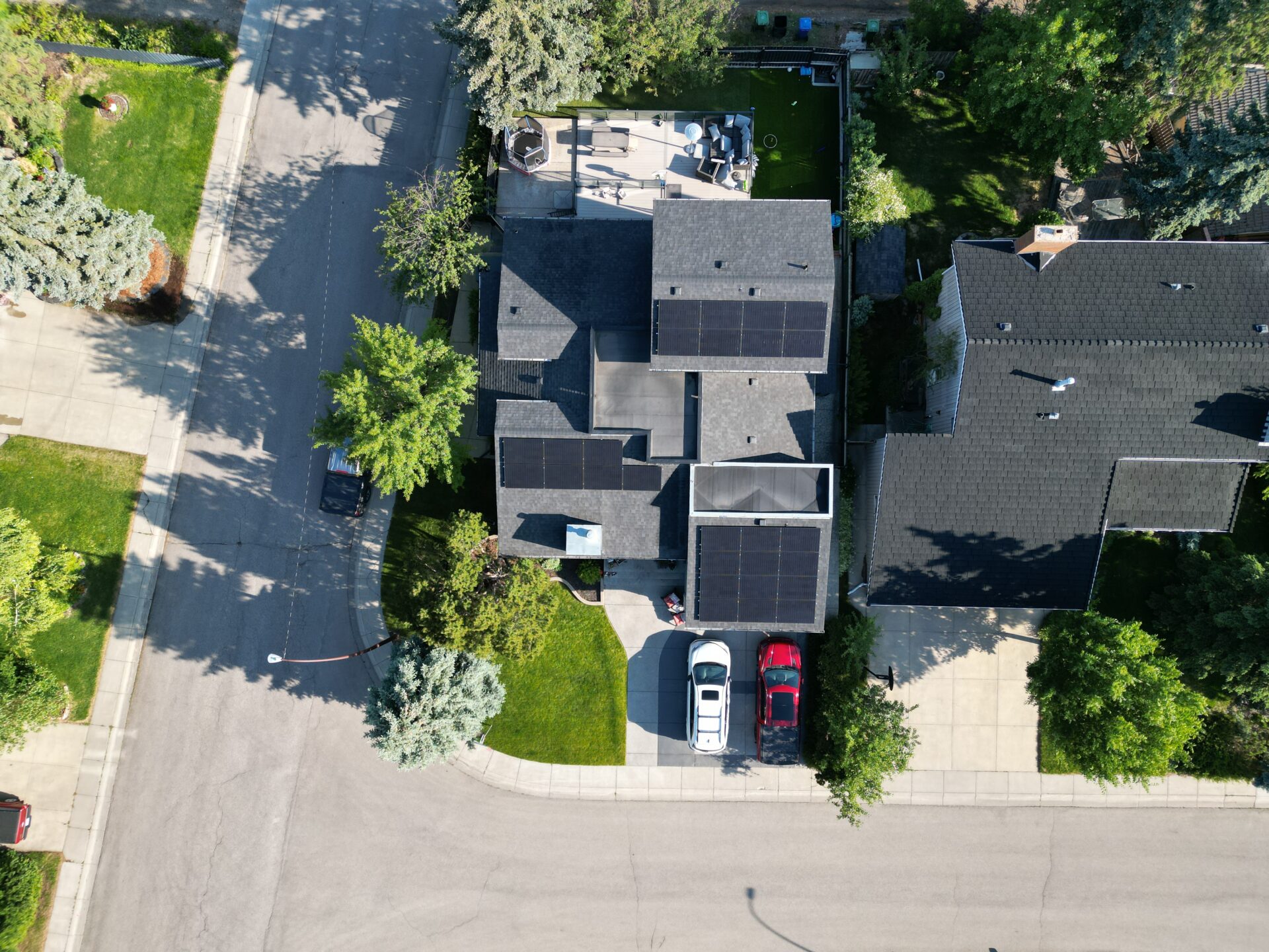 Aerial view of a residential area showing houses with solar panels, vehicles parked on driveways, and landscaped gardens with lush green trees.