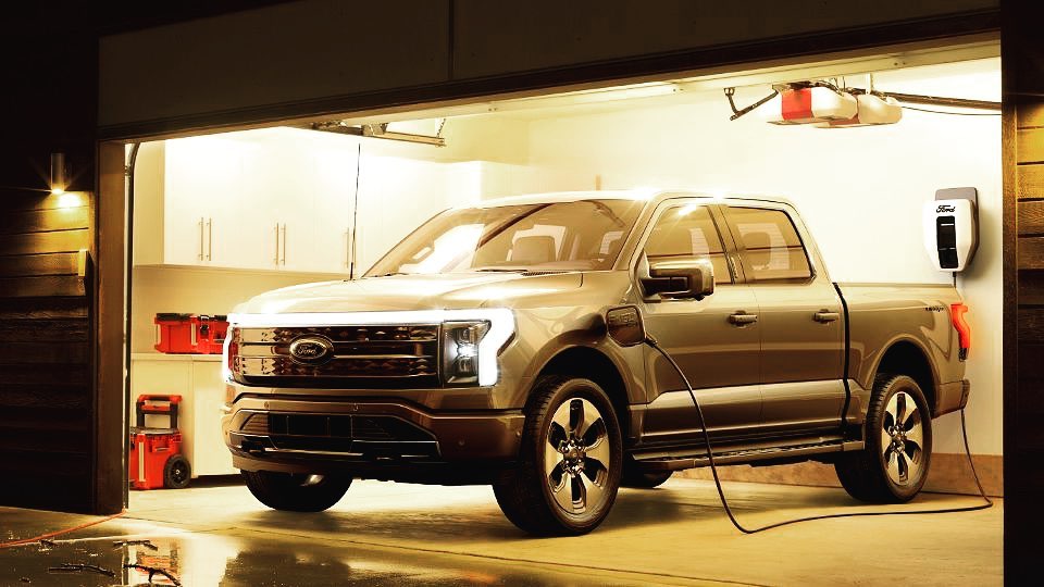 A modern pickup truck is charging in a well-lit garage with an EV charger mounted on the wall, indicating it's an electric vehicle.