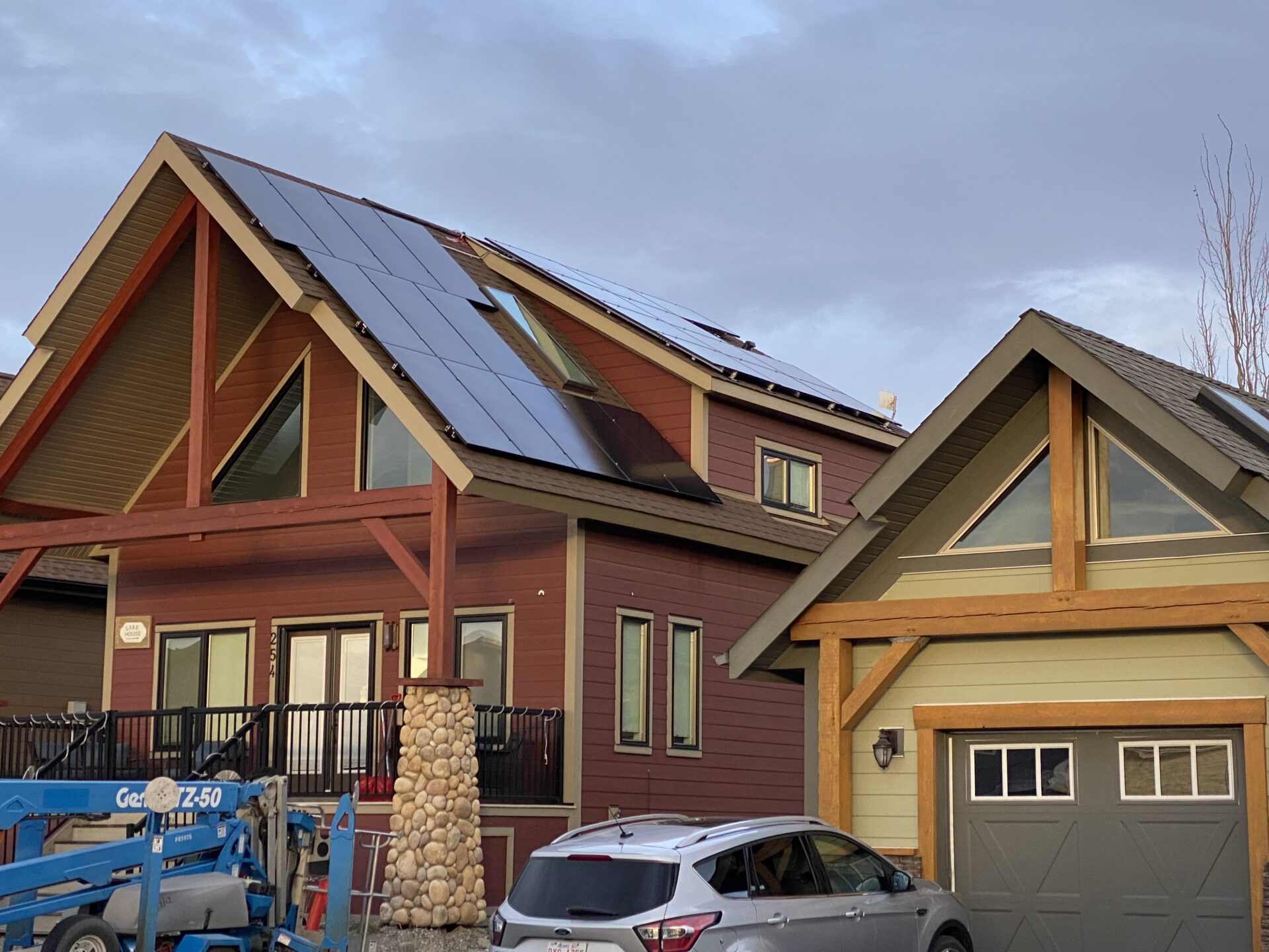 Two-story house with maroon and beige exterior, a slanted roof with solar panels, a balcony, and a stone column. Construction lift and vehicles in front.