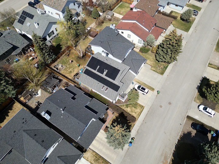 Aerial view of a suburban street with detached houses, manicured lawns, driveways with cars, and trees. Sunny day casting shadows on the ground.