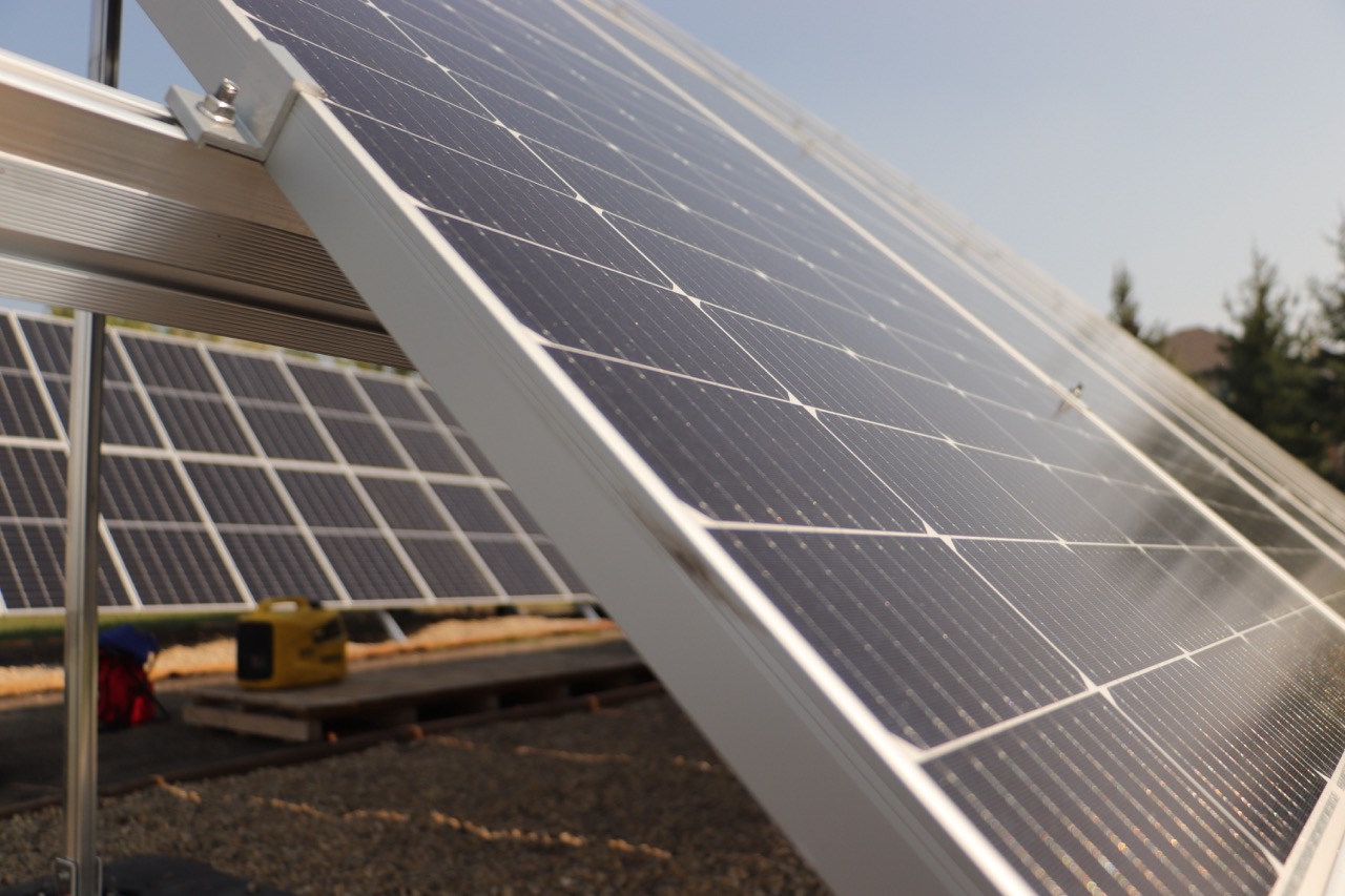 The image shows a close-up view of solar panels mounted on a metal framework, with a clear sky and trees in the background.