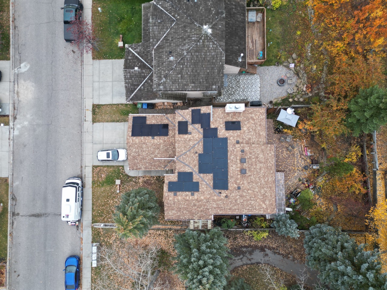 Aerial view of a residential area in autumn, showing houses with shingled roofs, vehicles, trees with fall foliage, and a neatly organized backyard.